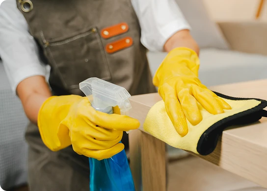 Pulizia di un tavolo in legno con un panno in microfibra e spray detergente indossando guanti protettivi.